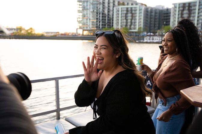 Two friends laughing on an urban riverboat deck at sunset, one waving and holding a canned drink with modern waterfront apartments in the background.