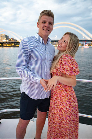 Smiling couple on a riverboat at dusk, woman in a pink floral dress showing an engagement ring while man in a striped shirt holds her hand, illuminated arched bridge and city lights reflected on the water behind them.