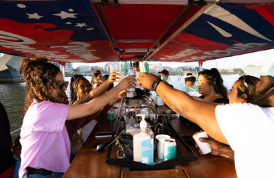 Friends toasting with cans and cups around a long bar on a covered party boat at an urban waterfront, city skyline and water visible, sunlight on faces and hand sanitizer on the counter.