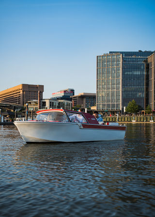 White-and-red motorboat cruising on an urban waterfront harbor with modern glass office buildings, sunlit calm water reflections, and a clear blue sky.