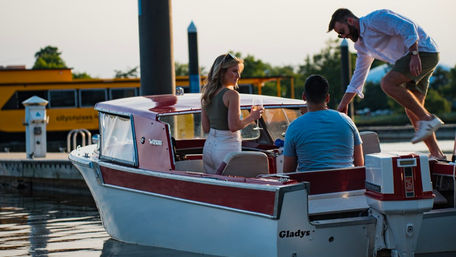 Three friends boarding a red-and-white motorboat at a marina at sunset, woman holding a wine glass while a man steps from the dock.