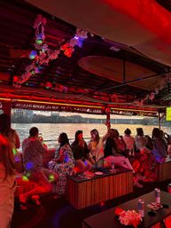 Red-lit tiki party boat on a river at sunset with a group enjoying drinks, surfboard ceiling decor, floral leis, and a bridge-lined city skyline visible through open sides.