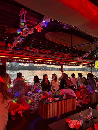Red-lit tiki party boat on a river at sunset with a group enjoying drinks, surfboard ceiling decor, floral leis, and a bridge-lined city skyline visible through open sides.