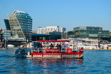 Red paddlewheel tour boat carrying passengers across a sunny urban marina, with docked yachts and modern glass waterfront buildings reflecting in blue water.