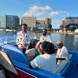 Four friends lounging on a blue speedboat at a sunny urban waterfront with modern glass buildings and city skyline in the background.