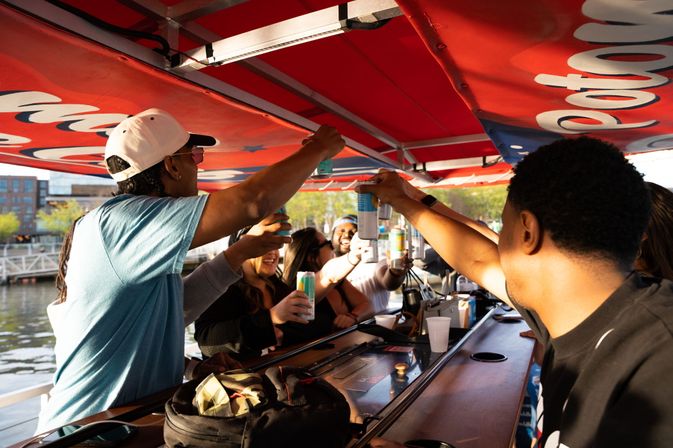 Sunlit group of friends toasting canned drinks at a covered outdoor bar on a boat, with an urban waterfront and dock visible in the background.