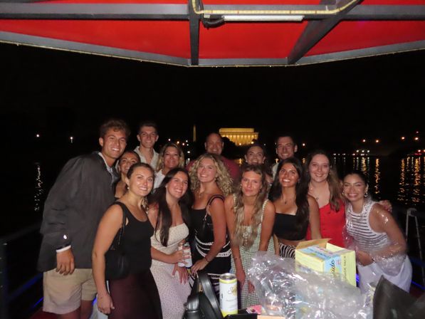 Smiling group of friends on a lively nighttime boat cruise on the Potomac River in Washington, D.C., with the illuminated Lincoln Memorial and Washington Monument reflected on the water