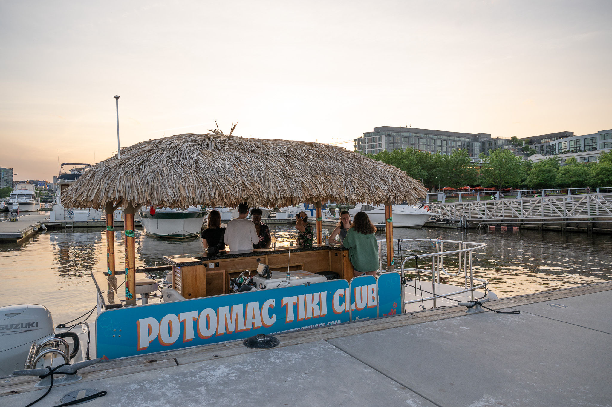 Thatched-roof tiki bar boat docked at a riverfront marina at sunset, with people chatting at the floating bar and yachts and waterfront buildings in the background.