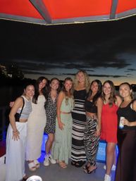 Smiling group of friends posing on a boat deck during a sunset cruise, with city skyline lights and water under a darkening sky.