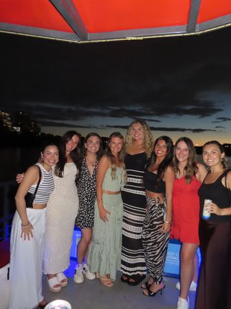 Smiling group of friends posing on a boat deck during a sunset cruise, with city skyline lights and water under a darkening sky.