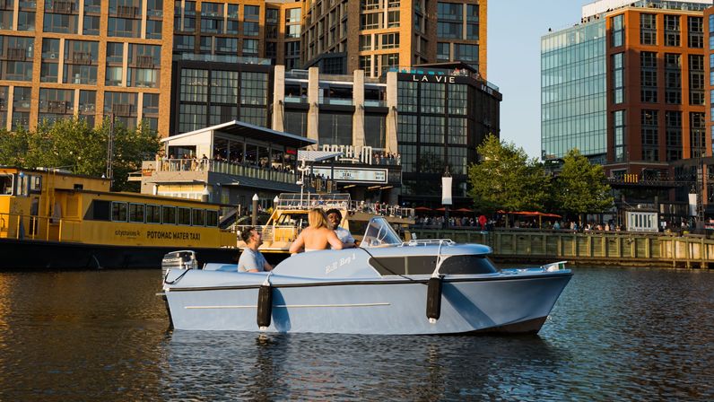 Three people on a light-blue motorboat cruising a sunny urban waterfront with a yellow water taxi nearby and modern mixed-use buildings along a busy river promenade.