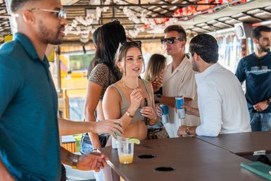 Young adults socializing at a sunny outdoor tiki-style bar, holding canned drinks and cocktails on the counter with a lively beachside vibe