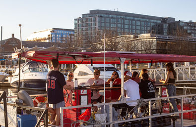 Friends enjoying a red pedal-powered party boat at a marina, docked beside yachts with modern waterfront apartments glowing in sunset light