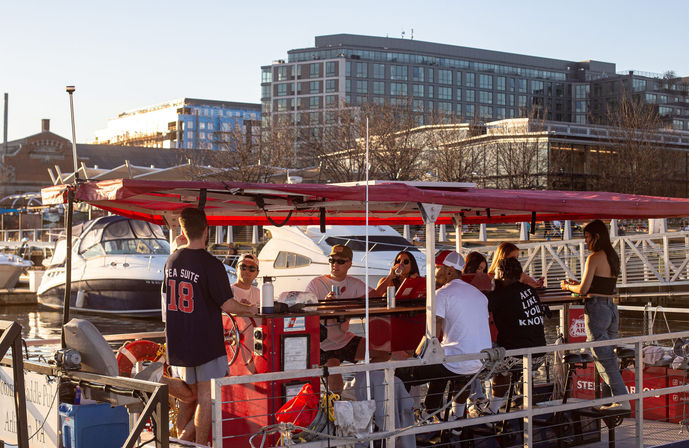 Friends enjoying a red pedal-powered party boat at a marina, docked beside yachts with modern waterfront apartments glowing in sunset light
