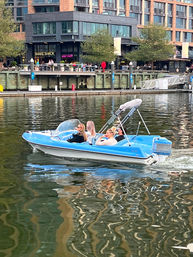 Blue recreational motorboat with four people cruising along a lively urban waterfront marina, passing restaurants, outdoor seating and modern apartment buildings reflected in the rippling water.