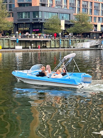 Blue recreational motorboat with four people cruising along a lively urban waterfront marina, passing restaurants, outdoor seating and modern apartment buildings reflected in the rippling water.