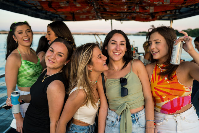 Group of friends in colorful summer outfits laughing and holding canned drinks on a sunset boat cruise over calm water with a distant shoreline.