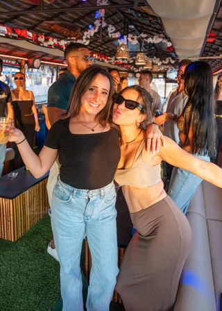 Two women posing and smiling at a lively sunset boat party — one holding a drink, the other wearing sunglasses and a crop top — surrounded by friends, tiki-style decor and warm golden light.