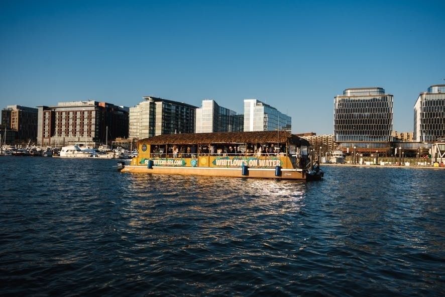 Sunlit tiki-style passenger boat cruising across an urban harbor with yachts and modern glass waterfront buildings lining the marina