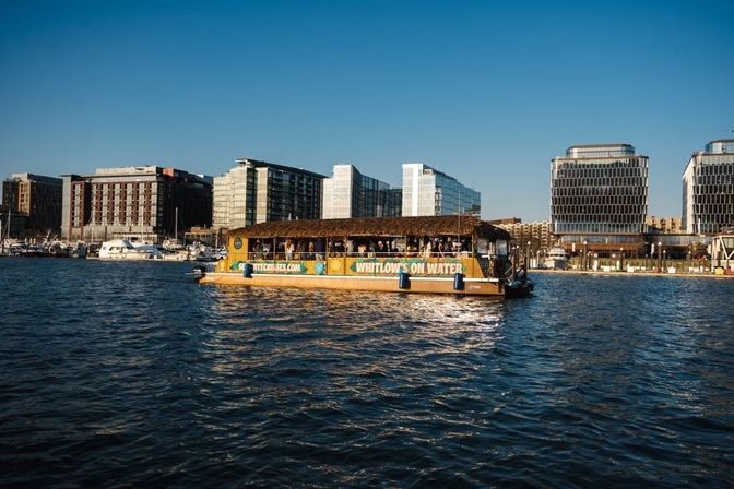 Sunlit tiki-style passenger boat cruising across an urban harbor with yachts and modern glass waterfront buildings lining the marina