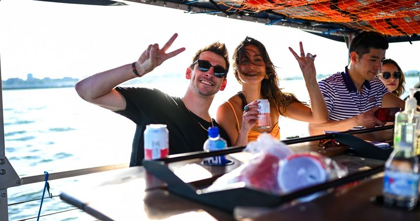 Friends enjoying a sunny boat cruise, smiling and flashing peace signs while holding canned drinks with open water and a distant shoreline in the background.