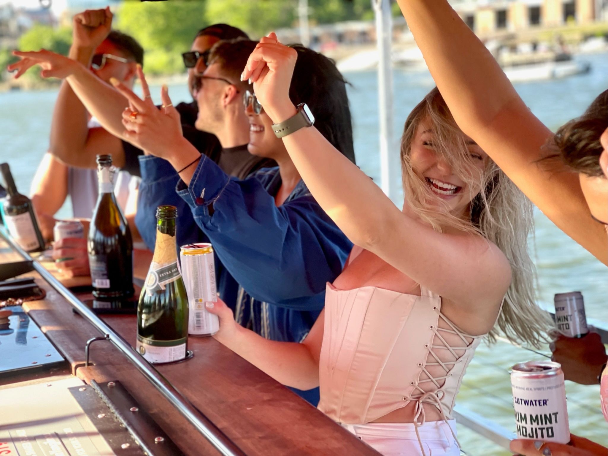 Smiling young woman in a corset laughing with friends at a sunny boat party by the waterfront, raising a canned cocktail at a wooden bar with champagne bottles nearby