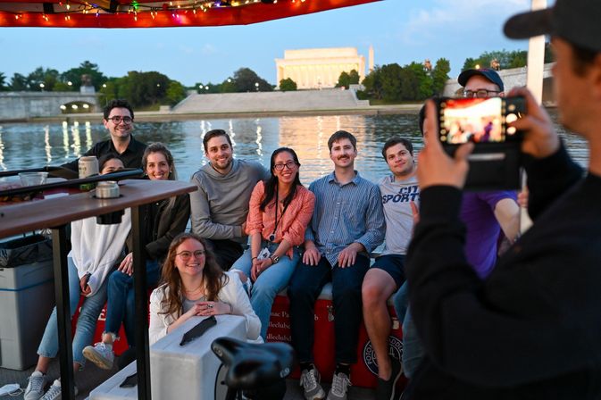 Smiling group on an evening boat cruise in Washington, D.C., posing for a photo with the illuminated Lincoln Memorial and Washington Monument reflecting on the water.
