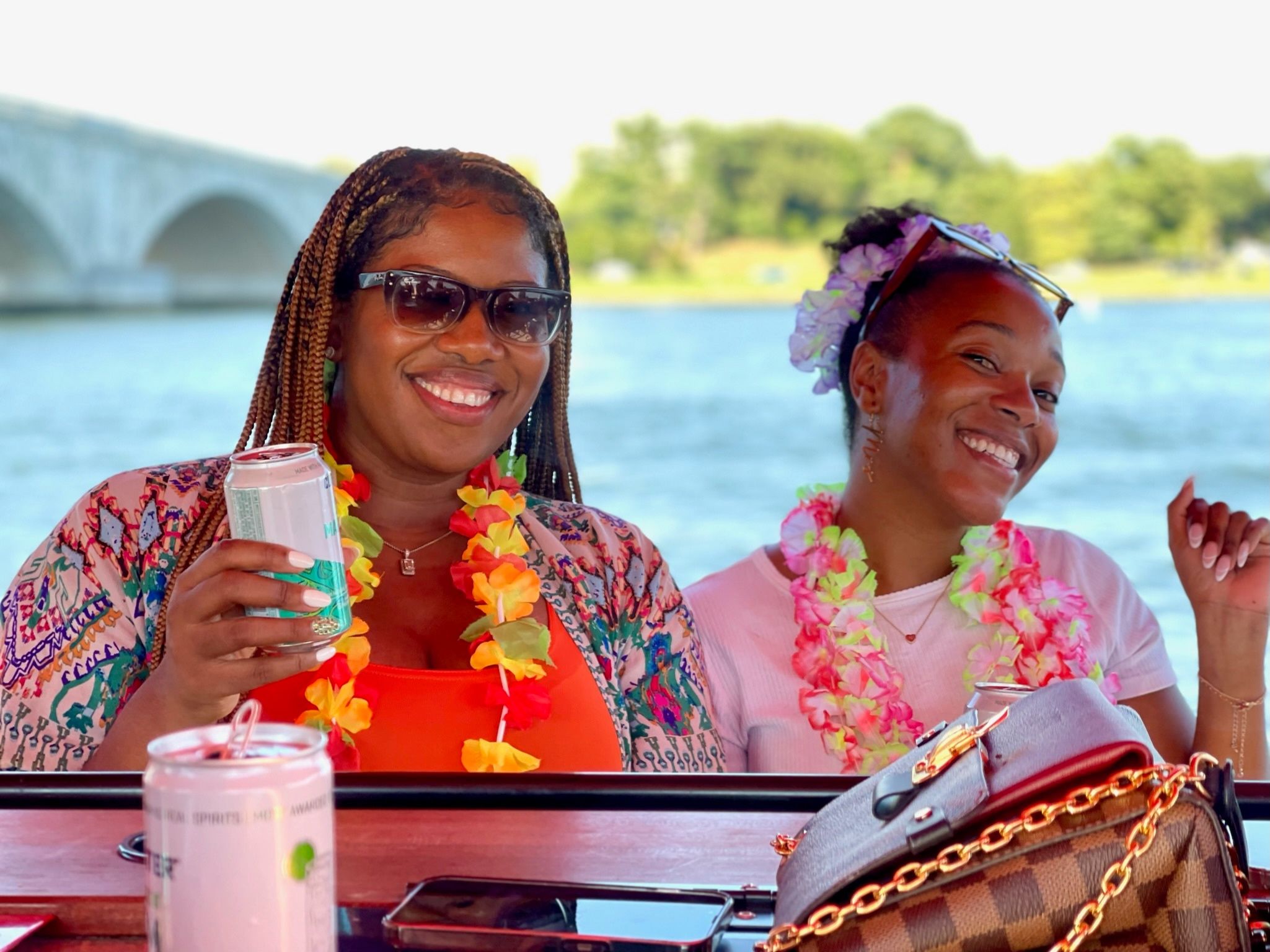 Two smiling friends in colorful leis and sunglasses enjoying a summer boat party on a river with a stone arch bridge in the background, holding canned drinks.