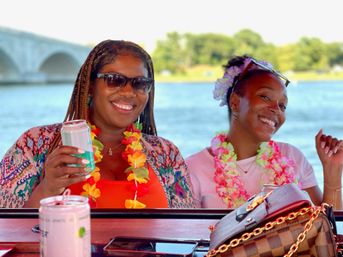 Two smiling friends in colorful leis and sunglasses enjoying a summer boat party on a river with a stone arch bridge in the background, holding canned drinks.