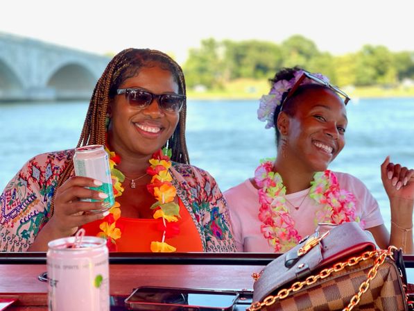 Two smiling friends in colorful leis and sunglasses enjoying a summer boat party on a river with a stone arch bridge in the background, holding canned drinks.