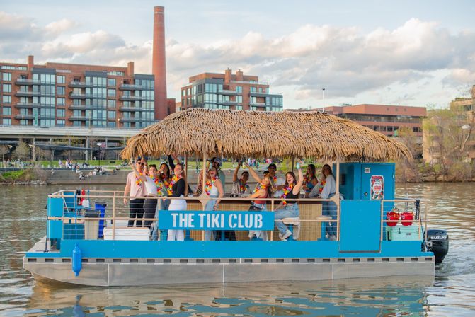 Group of people in colorful leis enjoying a tiki-style party boat with a thatched roof cruising a city riverfront past modern waterfront apartments.