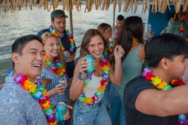 Group of young adults wearing colorful Hawaiian leis, laughing and holding canned drinks on a tiki-roofed party boat over calm water
