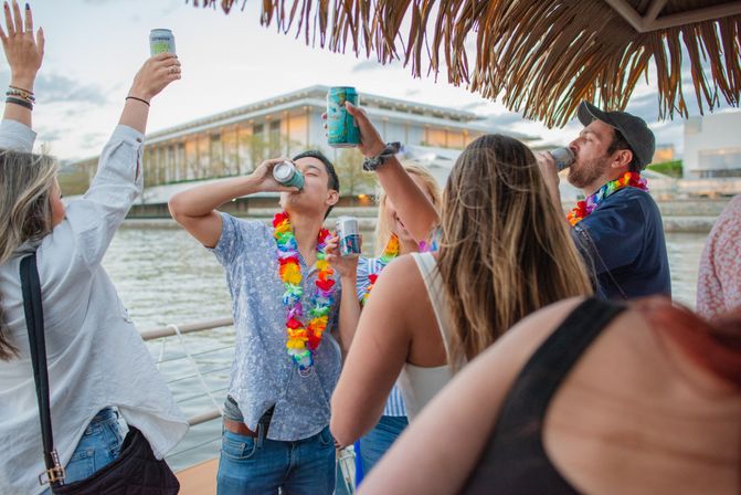 Group of friends on a waterfront boat party, wearing colorful leis and raising canned drinks under a thatched tiki roof during a river cruise by modern waterfront buildings