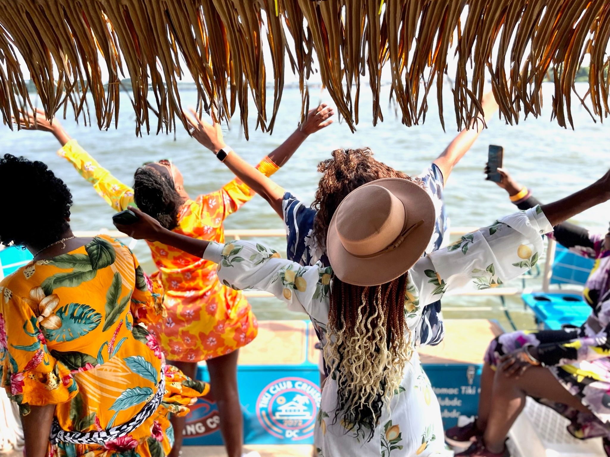Group of women in colorful tropical dresses and a sun hat cheering with arms raised on a thatched-roof boat, enjoying a sunny waterfront ride over calm coastal waters.
