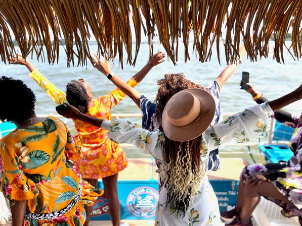 Group of women in colorful tropical dresses and a sun hat cheering with arms raised on a thatched-roof boat, enjoying a sunny waterfront ride over calm coastal waters.