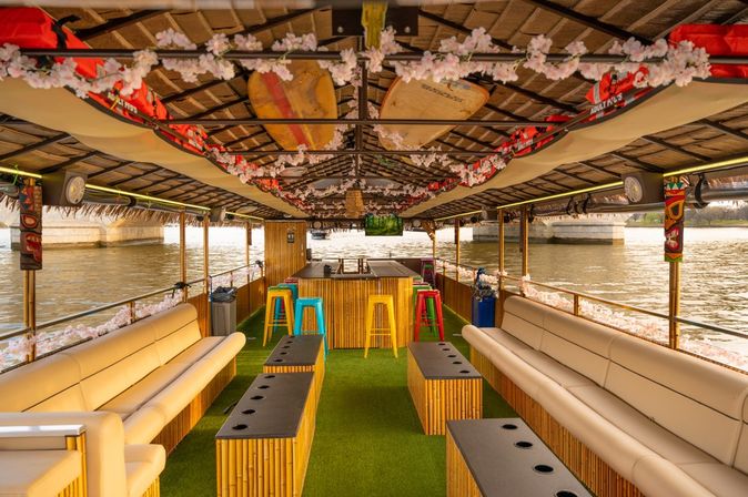 Bamboo-themed river party boat interior with cushioned lounges, tiki bar and colorful stools, cherry blossom garlands and lifejackets cruising under a bridge.