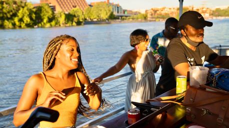 Group of friends on a sunset riverboat cruise along a city waterfront, smiling woman with braided hair dancing by the boat's outdoor bar