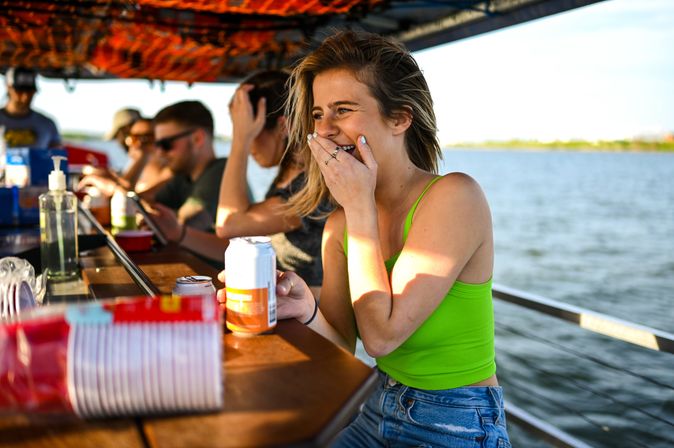 Young woman in a neon green tank top laughing and covering her mouth while holding a canned drink at a casual boat-side bar on a sunny lake, friends and drinks blurred in the background.
