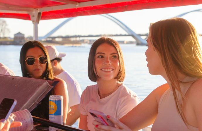 Three friends chatting and smiling on a small boat at a sunny urban waterfront with an arched bridge in the background; one wears sunglasses while others hold phones and a canned drink.