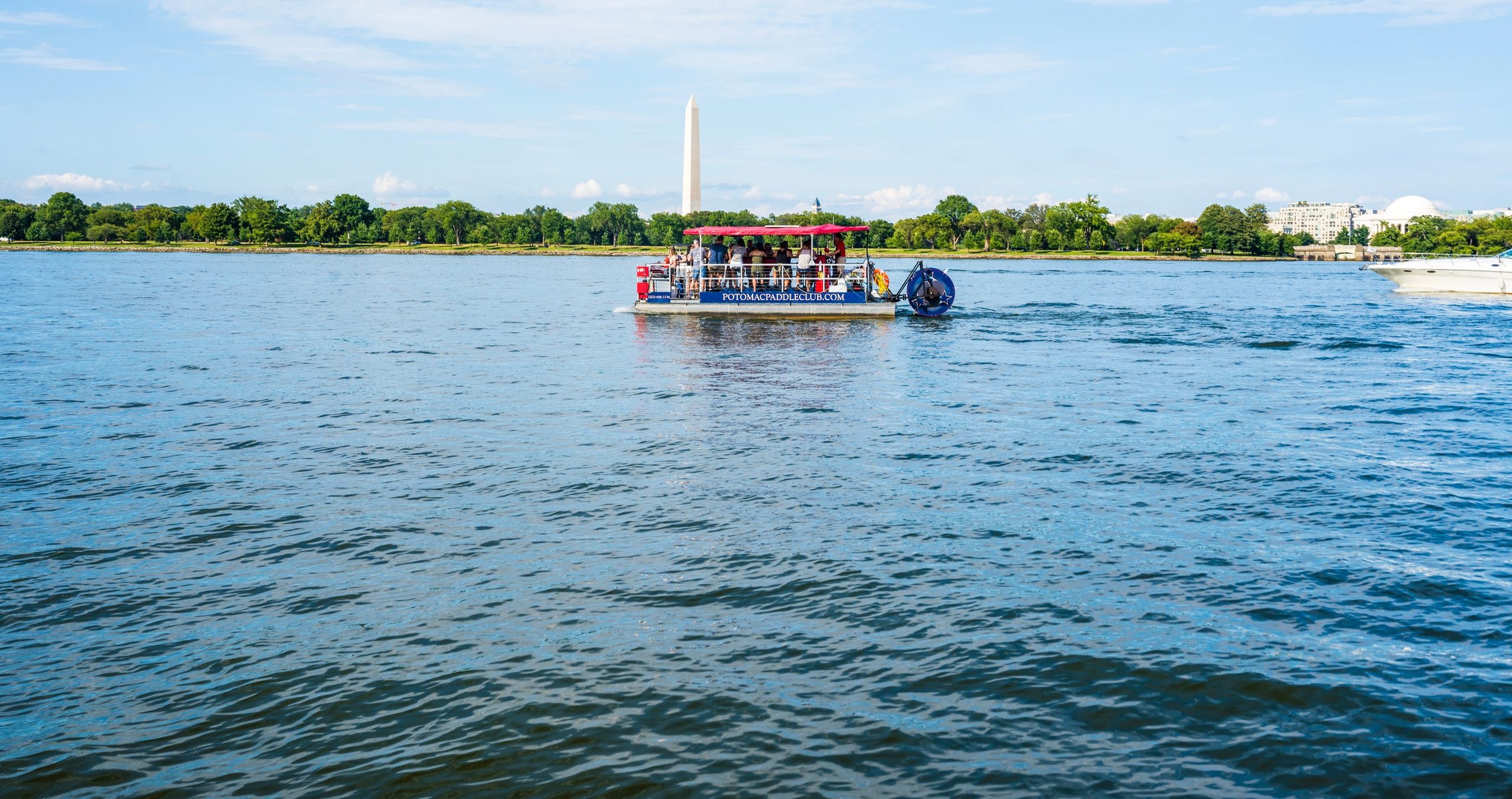 Pontoon tour boat on the Potomac River in Washington, D.C., cruising with the Washington Monument and tree-lined shoreline under a bright blue sky.