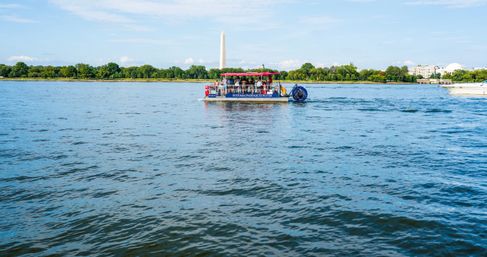 Pontoon tour boat on the Potomac River in Washington, D.C., cruising with the Washington Monument and tree-lined shoreline under a bright blue sky.