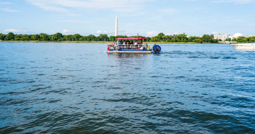 Pontoon tour boat on the Potomac River in Washington, D.C., cruising with the Washington Monument and tree-lined shoreline under a bright blue sky.