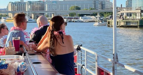 Group of people wearing pink leis on a sunny waterfront boat cruise with city skyline, bridge and docked boats in the background