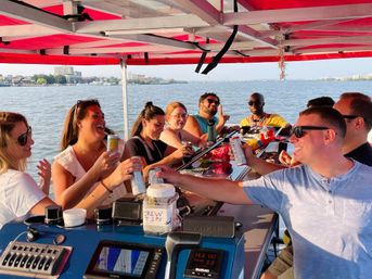Laughing group of friends cheering with canned drinks on a sunlit covered party boat, snacks and a visible "Crew Tips" jar on the bar with water and a distant waterfront skyline beyond.