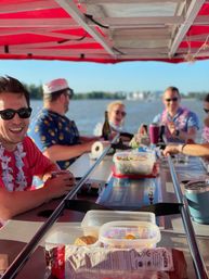 Friends on a covered party boat on a sunny lake, laughing around a central bar table with snacks, drinks, leis and sunglasses, shoreline blurred in the background.