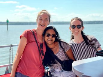 Three friends smiling on a sunny boat ride by the water — two in striped shirts and sunglasses and one in a red tee, waterfront and blue sky in the background.
