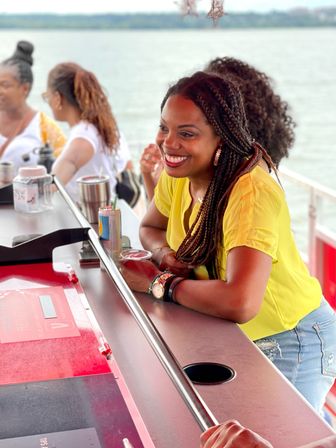 Smiling woman with braided hair in a yellow top leaning on a boat bar, enjoying drinks and laughing with friends on a sunny lake cruise