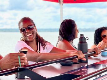 Smiling woman in sunglasses enjoying a lakeside boat bar on a summer cruise, friends leaning on the railing with a water bottle and phone
