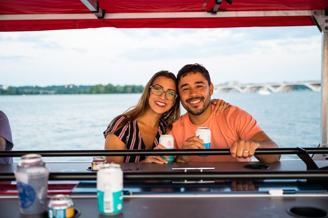 Smiling couple enjoying canned drinks on a covered riverboat, with waterfront trees and an arched bridge in the background.