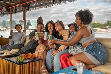 Group of friends laughing and clinking canned and mixed drinks aboard a covered riverboat, bamboo table with autumn décor and a city skyline across the water.